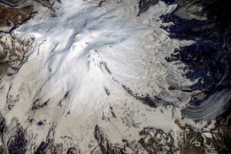 Рис. 13. На аэрофотоснимке Эльбруса хорошо видно наличие вулканических кратеров на двух вершинах (западный&nbsp;– крупнее, восточный&nbsp;– меньше). Черная нисходящая полоса справа по направлению «на 4 часа» - проступивший сквозь снег застывший лавовый поток&nbsp;[16]